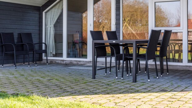 An outdoor patio with a table and chairs, a covered area with a sloped roof, and a view of trees through large windows. - Tranquil Cottage With Spa and Sauna-by Traum (Hemmet)