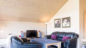 A modern living room with a wooden ceiling, a black leather sofa, a blue armchair, a coffee table, and a flat-screen TV. - Tranquil Cottage With Spa and Sauna-by Traum (Hemmet)