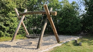 A wooden swing set with a tire swing, surrounded by sand and grass, with trees in the background. - Tranquil Cottage With Spa and Sauna-by Traum (Hemmet)
