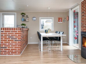 A modern kitchen with a brick accent wall, a dining area with a white table and black chairs, and a fireplace. - Holiday Home in FanÃ¸ (Fano)