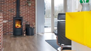A room with a brick wall, a wood-burning stove, a black leather chair, a yellow cabinet, and a wooden table. - Holiday Home in FanÃ¸ (Fano)