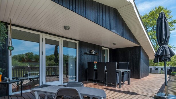 A covered outdoor area with a wooden deck, a table with chairs, and a glass door leading to a garden. - Holiday Home in FanÃ¸ (Fano)