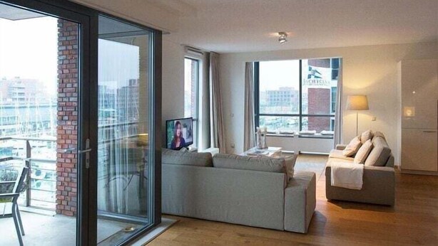 A modern living room with a large sliding glass door, a grey sofa, a small table, and a lamp. - Apartment in Scheveningen With Harbor View (The Hague)