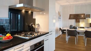 A modern kitchen with a black stove, white cabinets, and a dining area with a table and chairs. - Apartment in Scheveningen With Harbor View (The Hague)