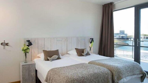 A modern hotel room with a large bed, a nightstand, and a view of the city through large windows. - Apartment in Scheveningen Near the Harbor (The Hague)