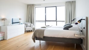 A modern bedroom with a large window, a bed with pillows, a bedside table with lamps, a chair, and a dresser. - Luxury Apartment With a View (The Hague)