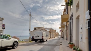 A narrow street with a white van parked on the side, buildings with balconies, and a clear view of the sea in the background.