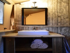 Een badkamer met een houten wastafelmeubel, een witte wasbak en een spiegel erboven. Handdoeken zitten opgeborgen in een houten kastje onder de wasbak.