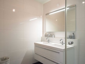 A modern bathroom with a white sink, two faucets, a mirror, and a tiled wall.