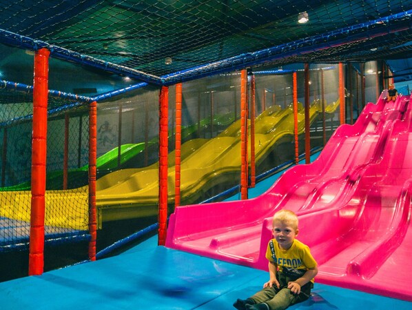 A child sitting on a blue slide in an indoor play area with multiple slides and netted walls.