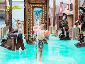 A child playing in a water fountain area with a pirate statue and a large door with a mural.