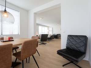 Un salon moderne avec un coin repas, comprenant une table en bois, des chaises marron, un canapé en cuir noir et une grande fenêtre avec des stores.