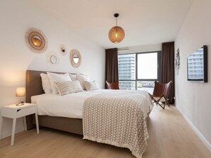 Ein modernes Hotelzimmer mit einem großen Bett, einem Nachttisch, einem Sessel und Blick auf eine Stadtlandschaft durch das Fenster.
