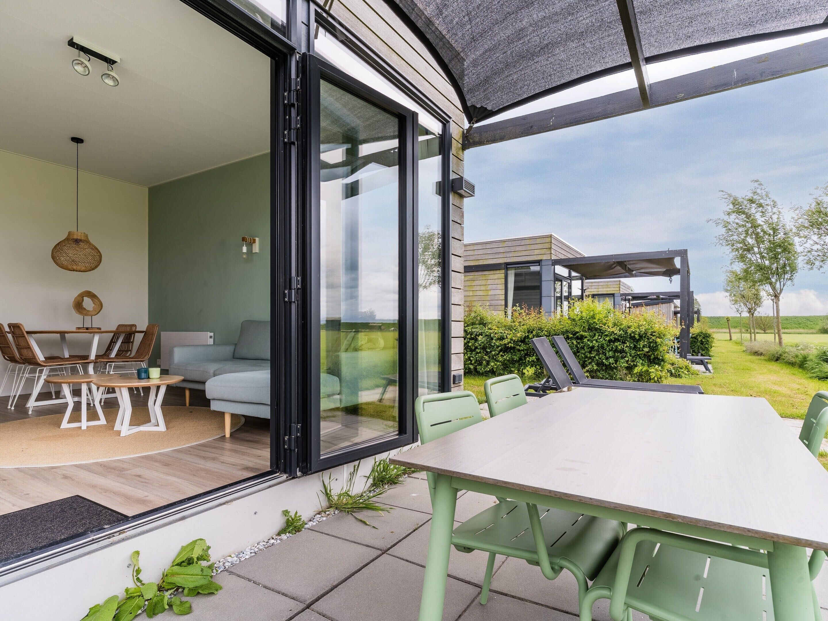 Un espacio exterior moderno para comer con mesa y sillas, una puerta corrediza de vidrio que da a un espacio interior, y una vista de una casa al fondo.