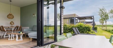 Un espacio exterior moderno para comer con mesa y sillas, una puerta corrediza de vidrio que da a un espacio interior, y una vista de una casa al fondo.