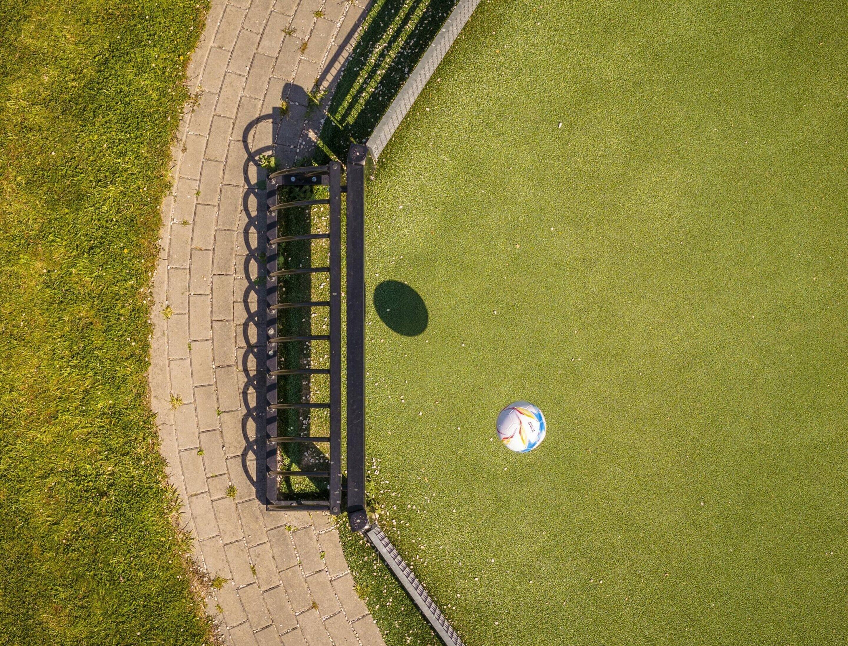 An aerial view of a park with a paved walkway, a metal fence, and a soccer ball on the grass.