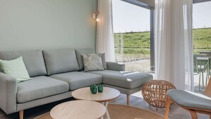 A modern living room with a grey sectional sofa, two round wooden tables, a wicker chair, and a view of a green landscape through large windows. - Lodge by Markermeer With Eco-friendly Design (Wijdenes)