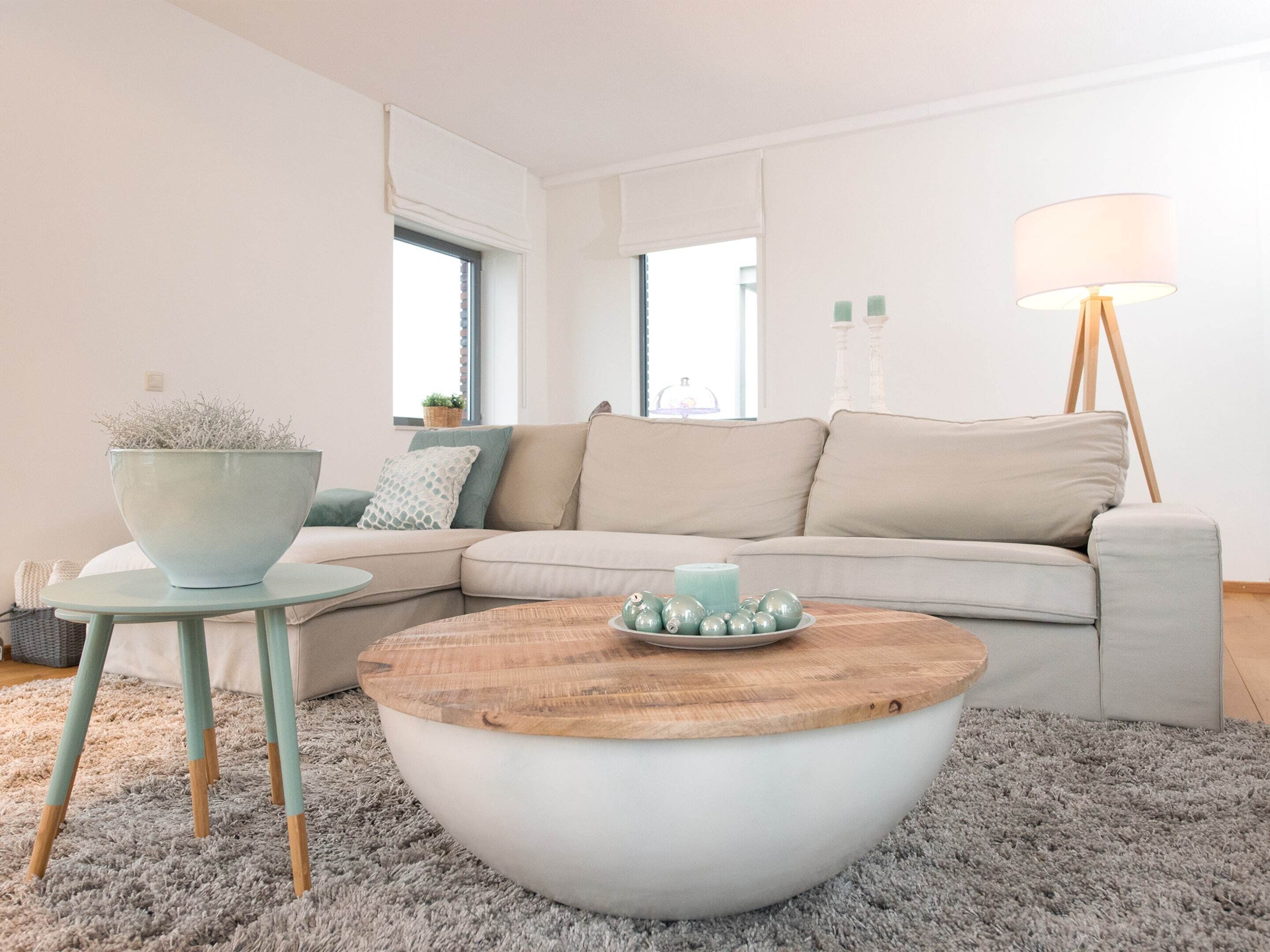 Un salon moderne avec un canapé d’angle beige, une table basse ronde en bois, une petite table d’appoint sur laquelle est posée une plante en pot et un lampadaire.