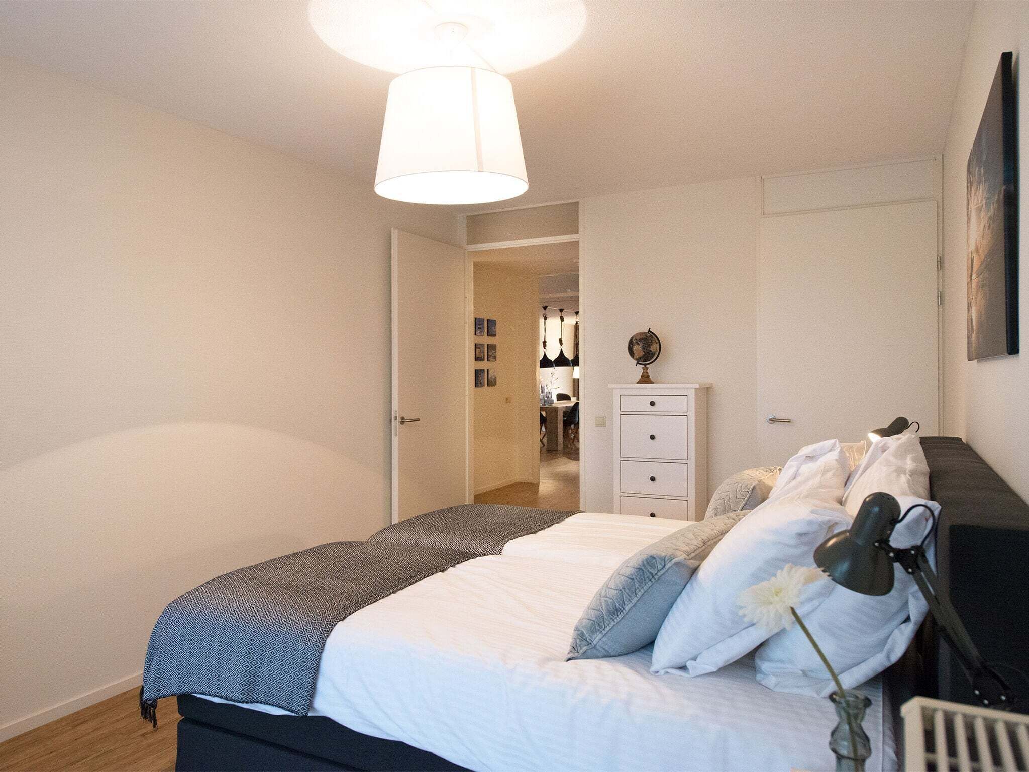 A bedroom with a bed, a nightstand, a lamp, a dresser, and a door leading to another room.