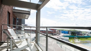 A balcony with a white chair, a glass railing, and a view of an industrial area with buildings and a road. - Luxury Apartment in Scheveningen Harbor (The Hague)