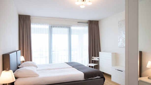 A modern hotel room with a large bed, a bedside table with a lamp, a chair, and a desk with a lamp. - Luxury Apartment in Scheveningen Harbor (The Hague)