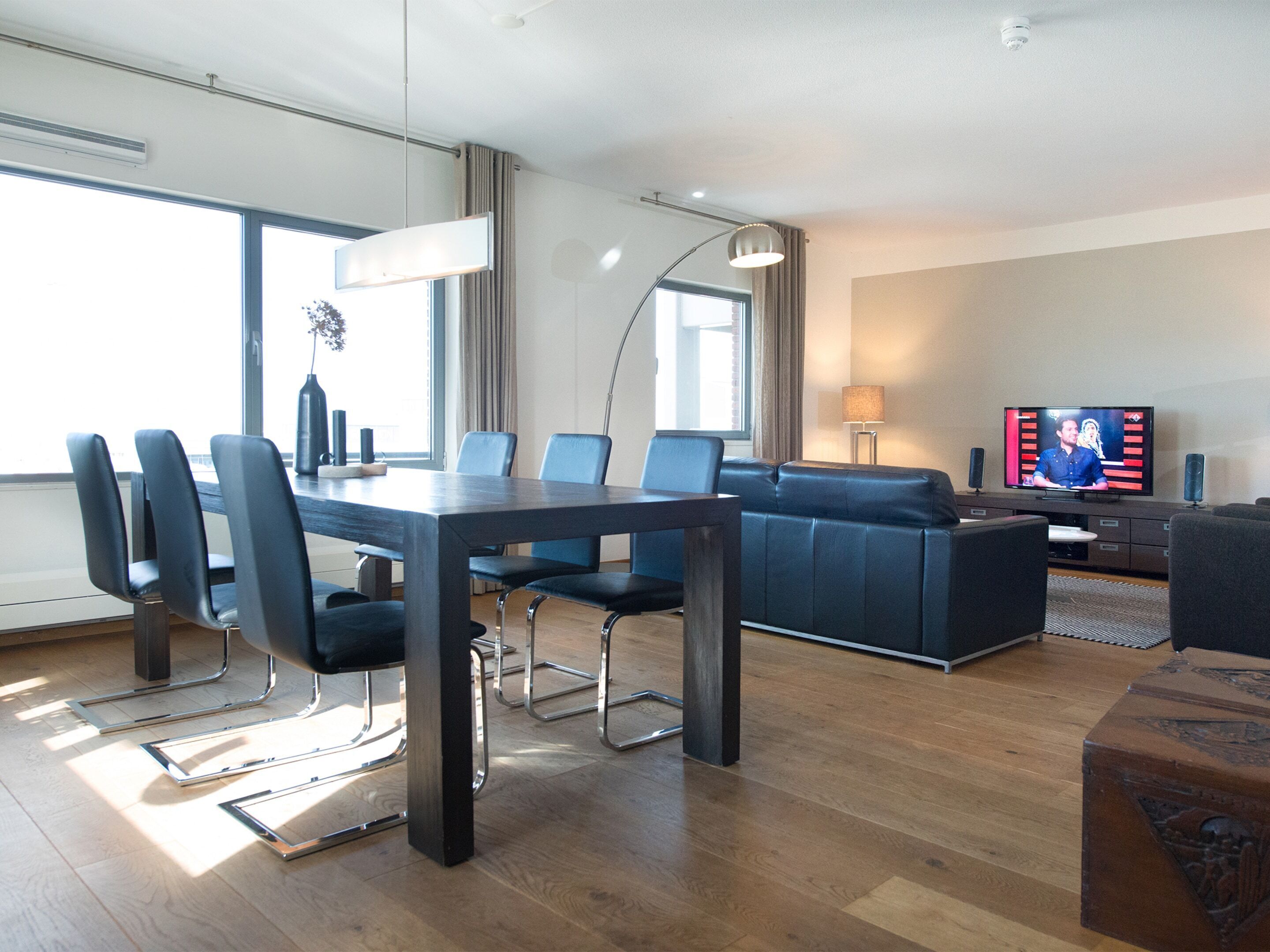 A modern hotel room with a dining area, a sofa, a television, and a large window.