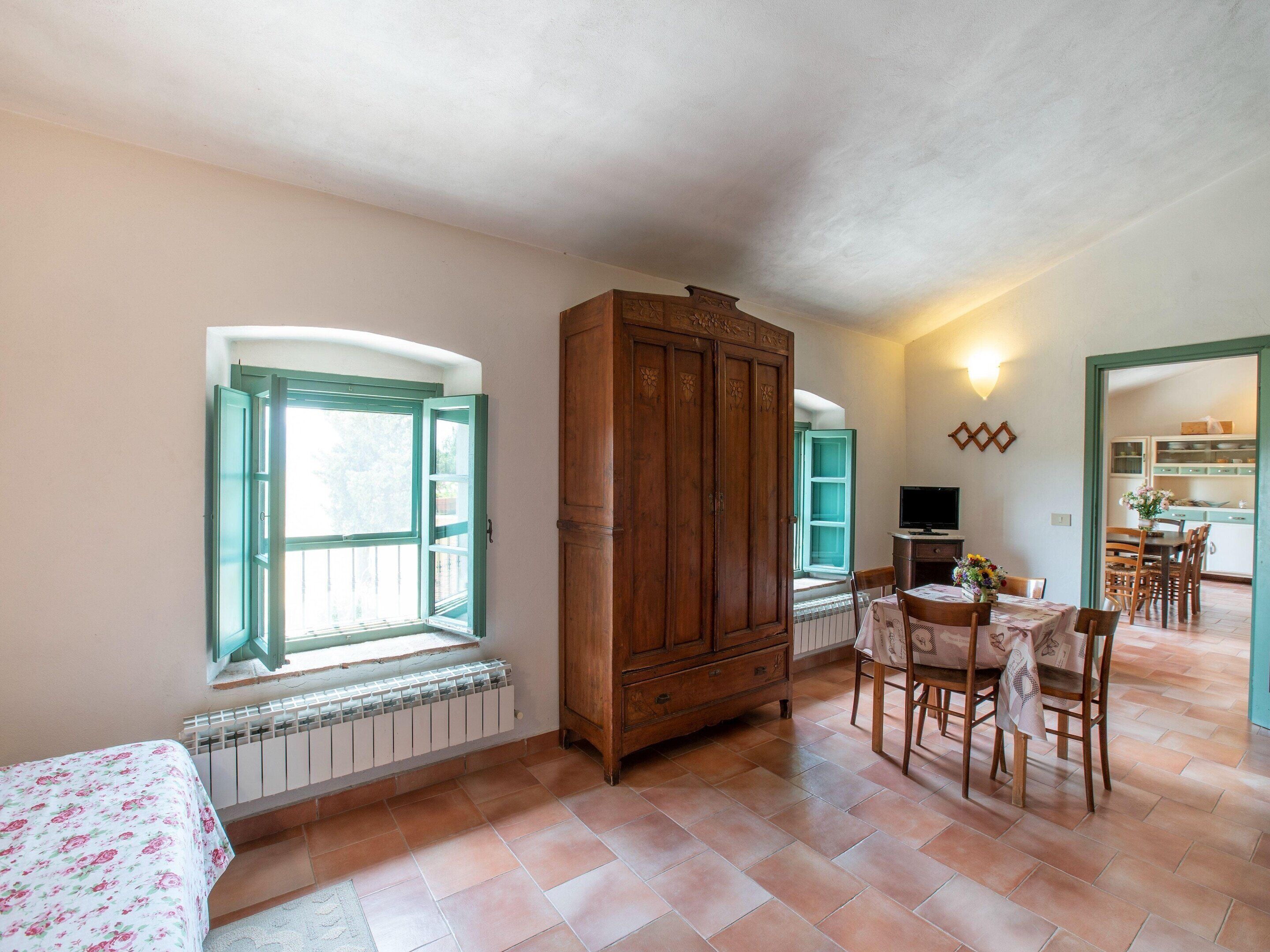 Una habitación con cama, un armario de madera, una mesa de comedor con sillas y una cocina visible a través de una puerta abierta.