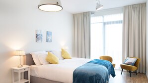 Une chambre d’hôtel moderne avec un grand lit, deux lampes de chevet, une petite table, une chaise et une grande fenêtre avec des rideaux.