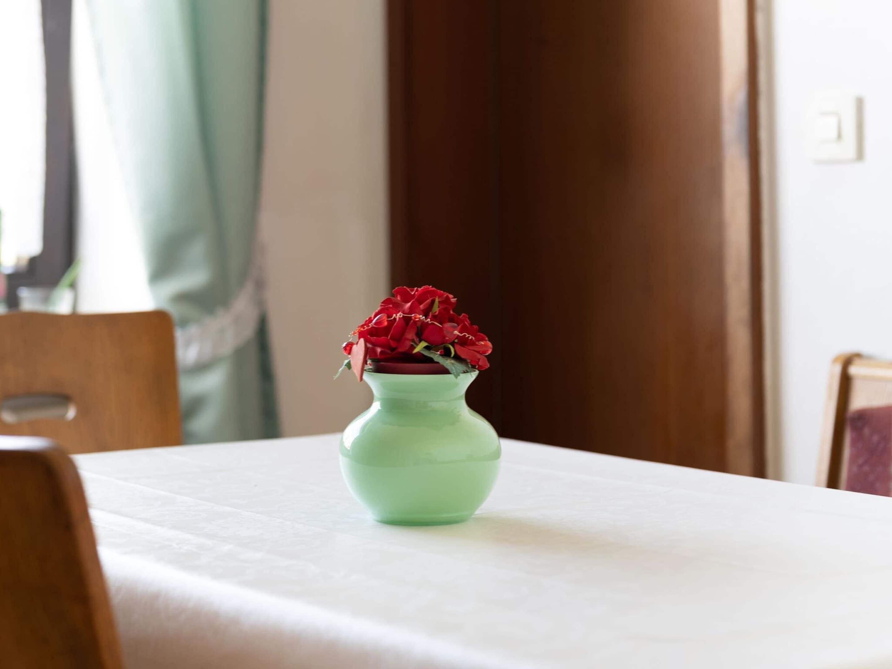 A table with a green vase containing red flowers, a wooden chair, and a white tablecloth.