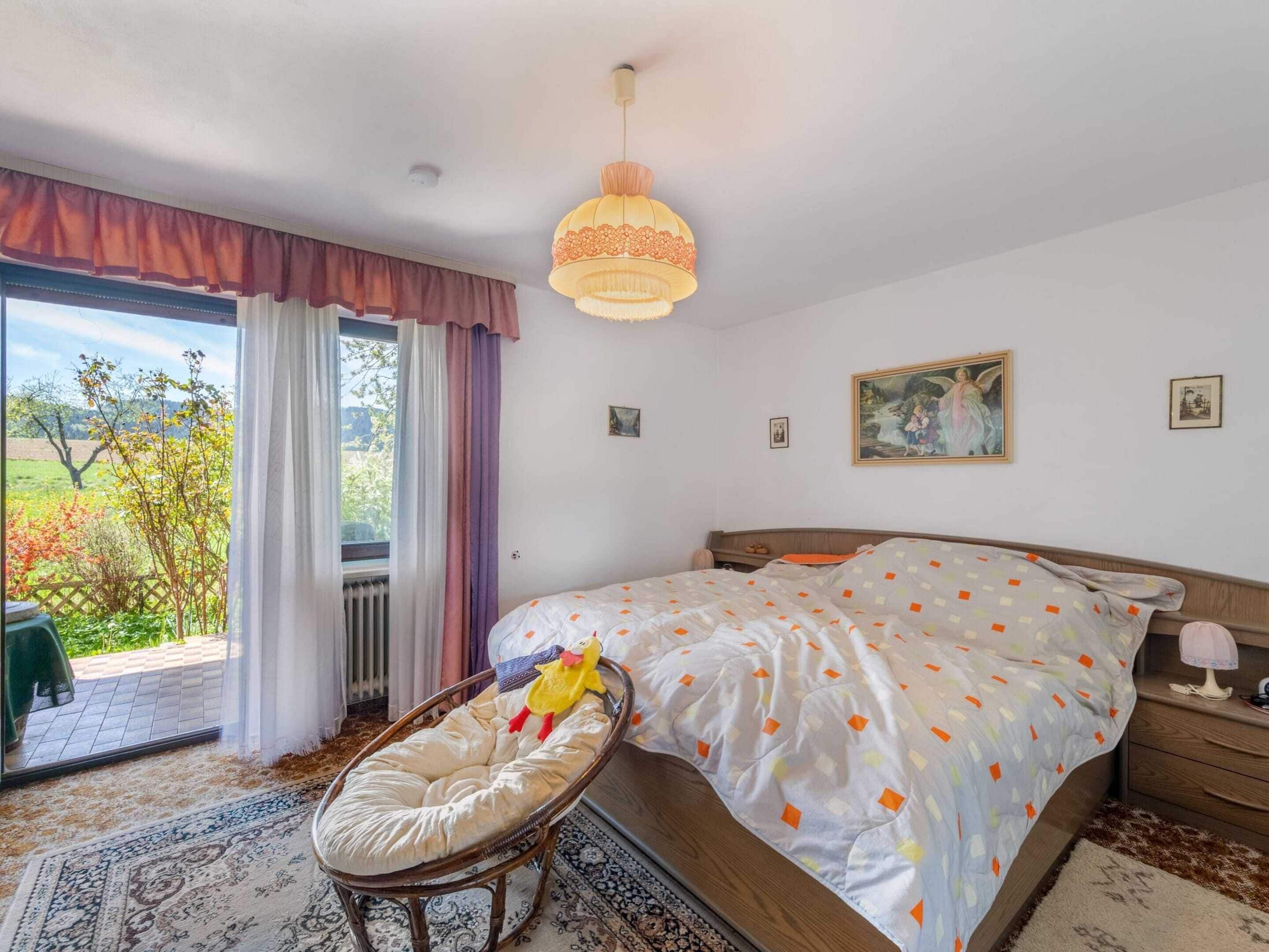 A bedroom with a large bed, a wicker chair, a hanging light fixture, and a view of a garden through the window.