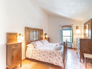Une chambre à coucher avec un lit en bois, deux tables de chevet, une commode, une chaise, une fenêtre avec des rideaux et un miroir.