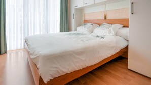Une chambre à coucher moderne avec un cadre de lit en bois, une literie blanche et une vue sur la ville à travers des rideaux transparents.