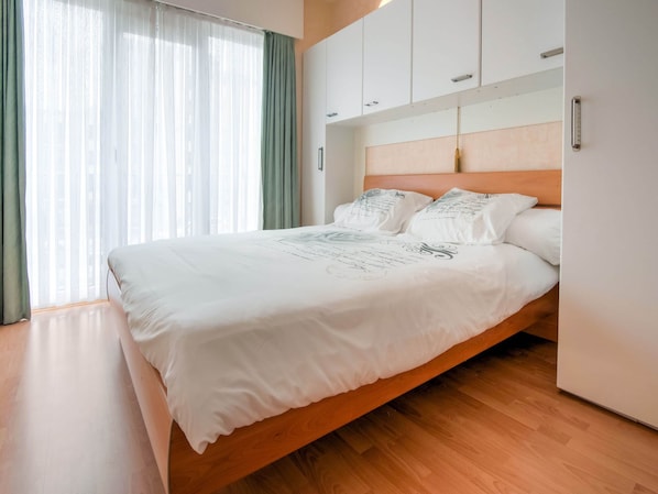 A modern bedroom with a wooden bed frame, white bedding, and a view of the city through sheer curtains.