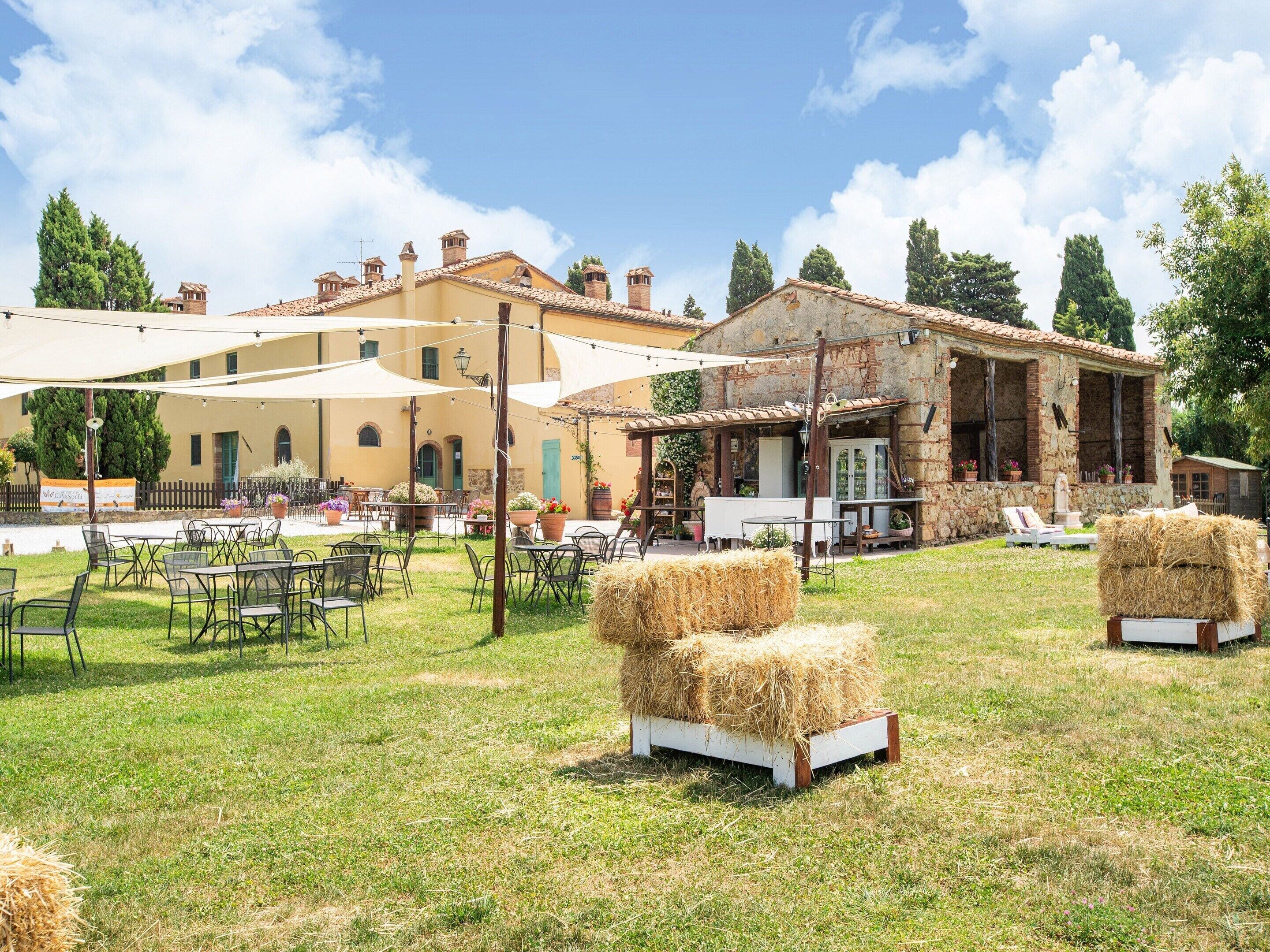 An outdoor seating area with tables and chairs, hay bales, and a building with stone walls and a tiled roof.
