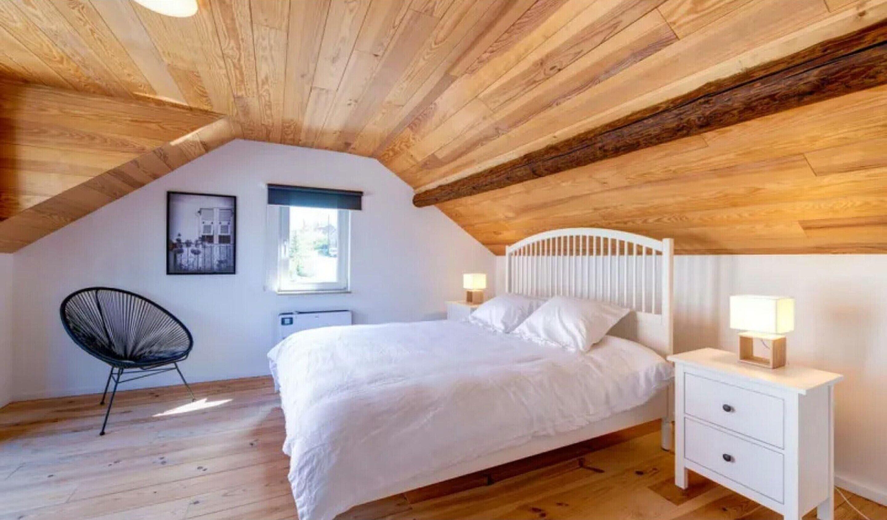 Une chambre avec un plafond en pente, un lit avec une literie blanche, une chaise, une table de chevet avec une lampe et une fenêtre.