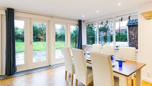 A dining area with a wooden table and white chairs, large windows, and a view of a garden. - Modern Holiday Home in Groede With a Terrace (Groede)