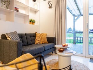 A modern living room with a grey sofa, a small round table, and a view of a wooden deck through a glass door.