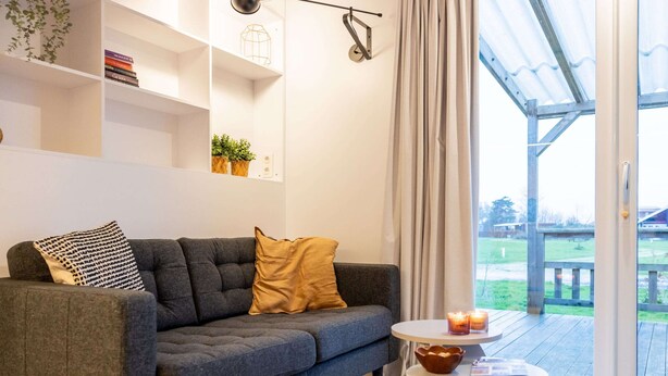 A modern living room with a grey sofa, a small round table, and a view of a wooden deck through a glass door.