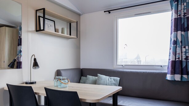 Een moderne kamer met een eettafel, stoelen, een bank, een raam met gordijnen en een plank met decoratieve objecten.