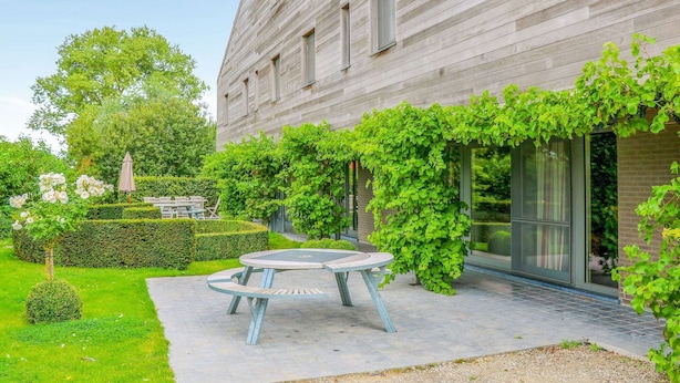 Un bâtiment moderne doté d’une façade en bois, d’un espace extérieur circulaire aménagé en coin salon et d’une végétation luxuriante.
