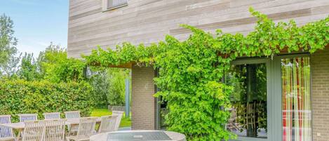 A modern house with a patio area featuring a round table and chairs, surrounded by lush greenery and a brick wall.