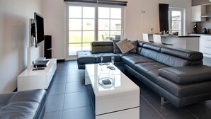 A modern living room with a sectional sofa, a white coffee table, and a flat-screen TV mounted on the wall. - Modern Holiday Home in De Haan With Garden (De Haan)
