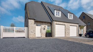 A two-story house with a garage, a white fence, and a gravel driveway. - Modern Holiday Home in De Haan With Garden (De Haan)