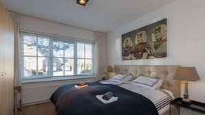 A hotel room with a large bed, a window with blinds, a painting on the wall, and a bedside table with a lamp. - Attractive Apartment near Center & Beach (Zoutelande)