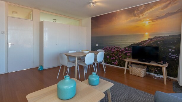 A modern living room with a dining area, a flat-screen TV, and a scenic sunset mural. - Romantic Coastal Retreat With Scenic Balcony (Zoutelande)