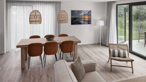 Un espace de repas moderne avec une table en bois, six chaises et un canapé. On y trouve une grande fenêtre avec des rideaux, une lampe fixée au mur et un tableau représentant un coucher de soleil.