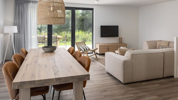 Un salon moderne avec une table à manger en bois, des chaises marron, un canapé beige et une grande fenêtre donnant sur un paysage verdoyant.