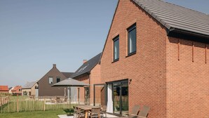 Une maison moderne en briques, dotée d’un toit à pente, d’une terrasse avec une table et des chaises, et d’une clôture en bois.