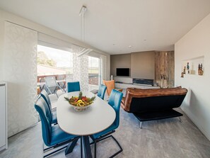 A modern living room with a dining table, blue chairs, a black sofa, a TV, and a large window.