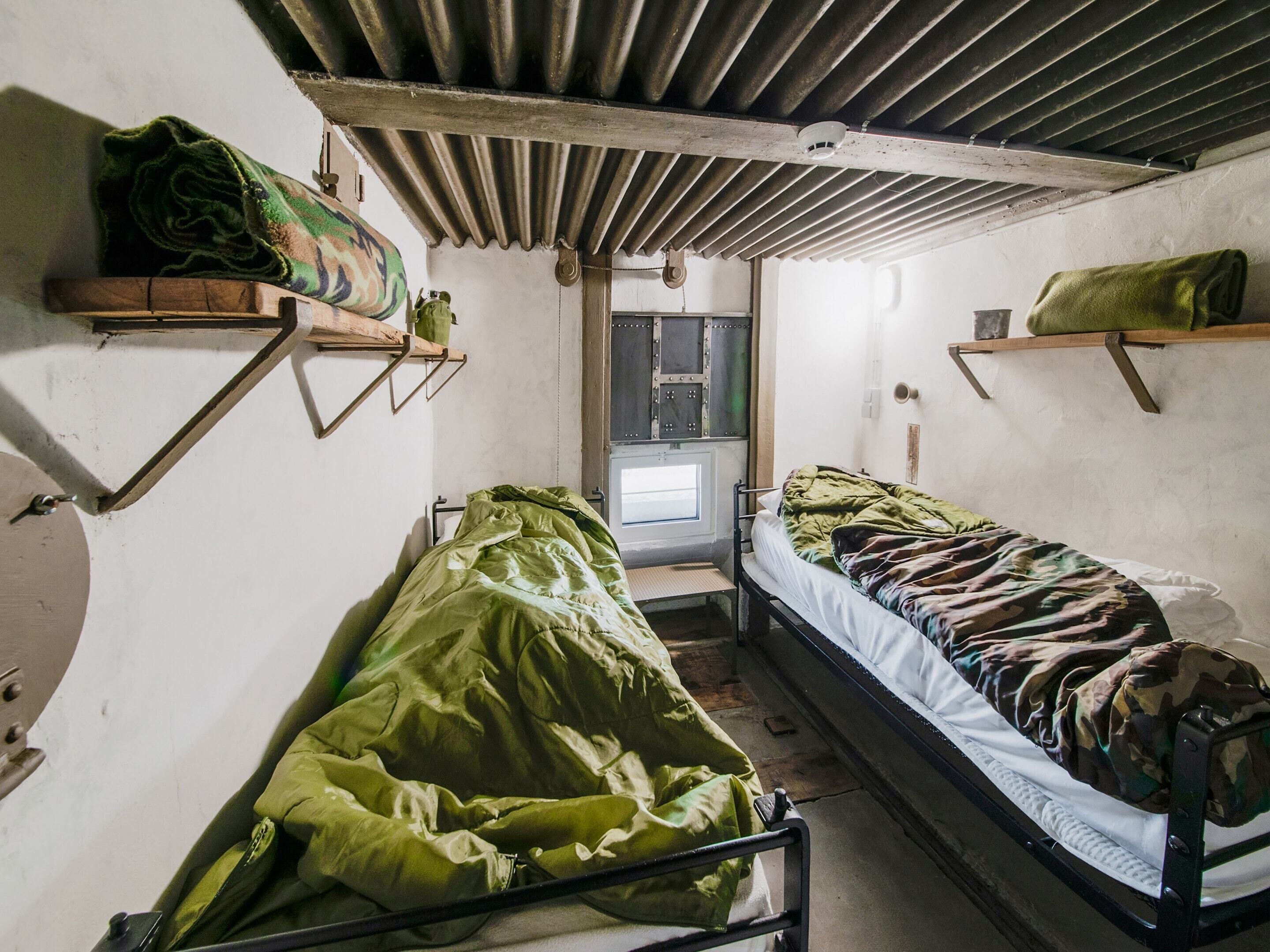 A small, dimly lit room with bunk beds, metal shelves, and a simple door.
